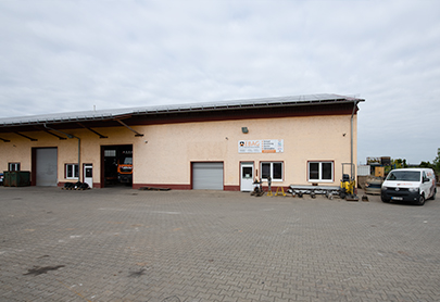 Ein breites, einstöckiges Industriegebäude mit beiger Fassade, mehreren Garagentoren und geparkten Fahrzeugen davor. Die Fläche ist mit Betonsteinen gepflastert und der Himmel ist bewölkt.